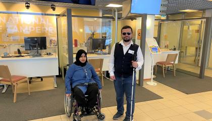 Liikuntavammainen nainen istuu pyörätuolissa. Hänen oikealla puolellaan seisoo näkövammainen mies, jolla on kädessä valkoinen sokean keppi. Molempien kasvot ovat kohti kameraa.