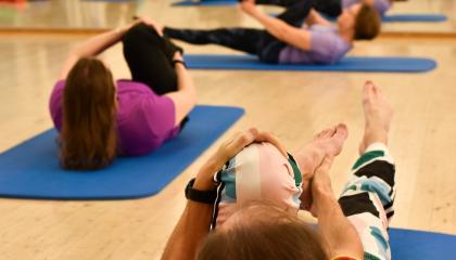 Kolme henkilöä harjoittaa pilatesta lattialla jumppamatoilla maaten. He pitävät toisesta polvestaan kiinni.