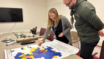 Nainen ja mies ovat kumartuneet pöydän ylle tarkastelemaan käsitekarttajulistetta kokoushuoneessa.