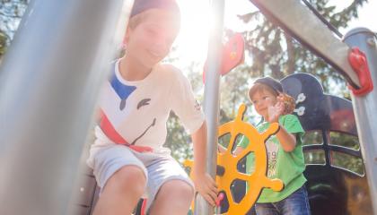 Kaksi lasta leikkii ulkona laivaa muistuttavalla leikkitelineellä. Vihreäpaitainen poika ohjaa keltaista ruoria, valkopaitainen poika arpoo, laskeutuuko metallista tankoa pitkin alas maahan.