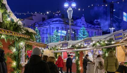 Ihmisiä hämärän aikaan Joulutorilla joulukoristelujen ja valaistujen puotimökkien äärellä, taustalla valotaiteella valaistu rakennus.