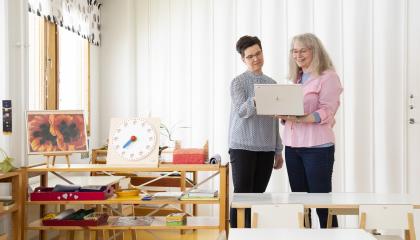 Kaksi naista katsoo seisten jotain kannettavalta tietokoneelta ja hymyilevät. Etualalla valkoisia pöytiä, naisten vasemmalla puolella matala hylly, jossa sekalaista tavaraa, kuten lasten opetuskello.