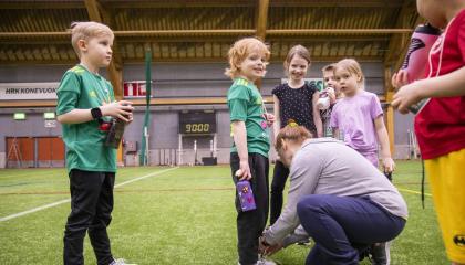 Lapsia rivissä urheiluvaatteissa nurmikentällä. Aikuinen sitoo yhden lapsen kengännauhoja.