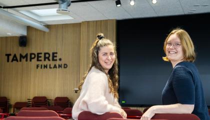 Kaksi henkilöä istuu punaisilla tuoleilla. He ovat kääntäneet katseensa kuvaajaan päin. Taustalla on teksti Tampere.Finland.