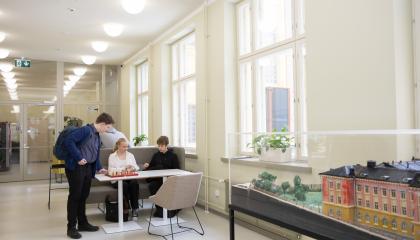 Kolme nuorta juttelee keskenään pöydän ääressä. Kuvassa oikealla on koulurakennuksen pienoismalli. 