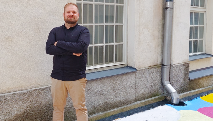 Mies seisoo kädet puuskassa vaalean rakennuksen seinämällä, taustalla ruutuikkuna.