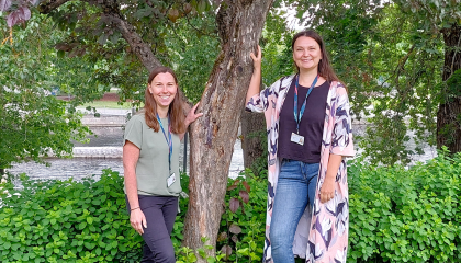 Kaksi hymyilevää naista nojaa puuhun, yksi kummallakin puolella.