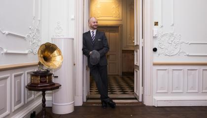 Henkilö seisoo hymyillen nojaten oven pieleen. Oven vasemmalla puolella katsojasta nähden on pienellä pöydällä gramofoni.