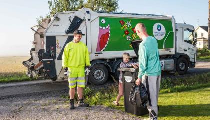 Jäteauto noutaa jätteet jäteastiasta. Etualalla kuljettaja ja mies ja poika, joilla mukana jätesäkki.