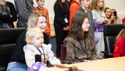 Hymyilevä äiti istuu pöydän ääressä pikkulapsi sylissään. Pöydän ääressä ja takana on myös muita lapsia.