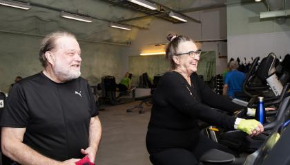 nainen polkee kuntopyörällä, mies katsoo vieressä.