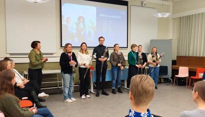 Sertifioitujen kukittamistilaisuudessa otetun kuvan keskellä sertifioidut ihmiset rivissä, yleisön ympäröimänä.