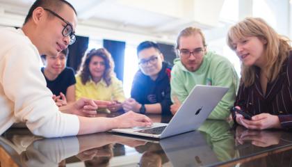 Kuusi henkilöä iloisesti katsomassa tietokoneen näyttöä valoisassa toimistotilassa.