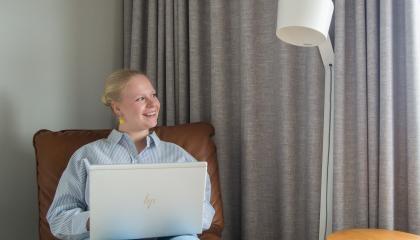 Vaaleatukkainen henkilön istuu tuolilla ja katsoo iloisesti sivulle. Taustalla näkyy harmaat verhot ja lattialamppu.