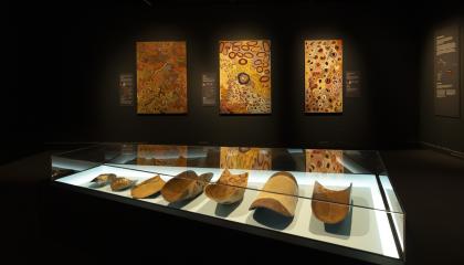 Hämärässä näyttelytilassa on seinillä värikkäitä maalauksia. Edessä lasivitriinissä puisia esineitä.