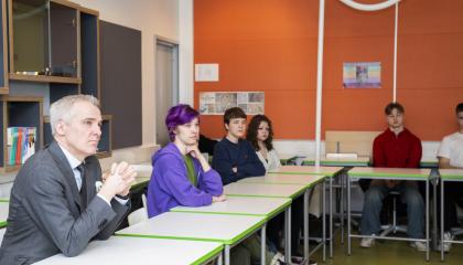 Ministeri ja opiskelijat istuvat rinnakkain luokkatilassa, jonka yksi seinä on voimakkaan oranssin värinen.