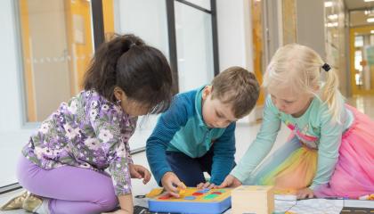 Kolme päiväkoti-ikäistä lasta istuu lattialla ison ikkunan edessä ja leikkii palikoilla.