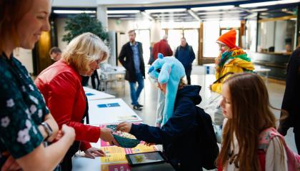 Kaupungin työntekijä tarjoaa esittelypöydän ylitse makeisia lapselle. Lapsella on päässään hauska vaaleansininen eläintä esittävä lakki. Taustalla näkyy muitakin lapsia ja aikuisia.