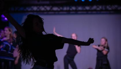 Pitkähiuksinen henkilö tanssii kuvan etualalla. Taustalla epätarkkana muita tanssijoita.