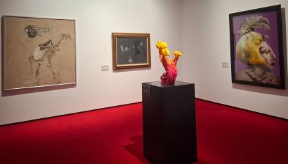 Näyttelytila, jossa on valkoiset seinät ja punainen matto. Seinillä kolme maalausta ja edessä oranssi kukkamainen veistos. 