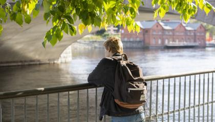 Yksi seisova nuori nojaa sillan alla kaiteeseen ja katsoo virtaavaa vettä. Nuoren pään päällä roikkuu puunoksia lehtineen. Vastarannalla näkyy punatiilinen rakennus. 