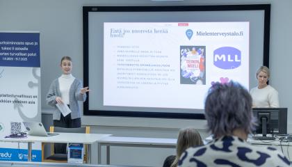 Kaksi henkilöä seisoo luentotilan etuosassa ja taustalle on heijastettu kuva tilaisuuden aiheesta.