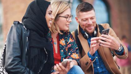 Kolme hymyilevää ihmistä istuu rinnakkain. Yksi heistä näyttää muille jotakin puhelimen näytöltä.