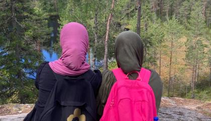 Kaksi nuorta henkilöä istuu vierekkäin korkealla kalliolla ja katselee metsäistä maisemaa, jossa näkyy myös pieni lampi. 