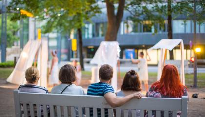Viisi henkilöä istuu Hämeenpuistossa penkillä ja katsoo edessä olevaa taide-esitystä.