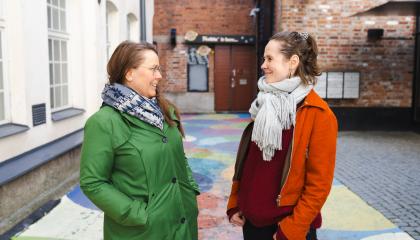 Kaksi hymyilevää naista katsoo toisiaan. Toisella on vihreä ja toisella oranssi takki.