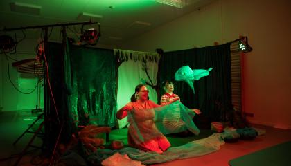 Käynnissä on Metsäretki-niminen esitys, jossa vihreät lavasteet ja kaksi esiintyjää heiluttelee sinisiä huiveja. 