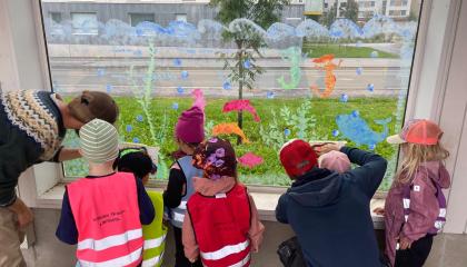 Kuusi värikkäisiin huomioliiveihin pukeutunutta lasta piirtää ikkunaan ikkunatusseilla yhdessä kahden aikuisen kanssa.