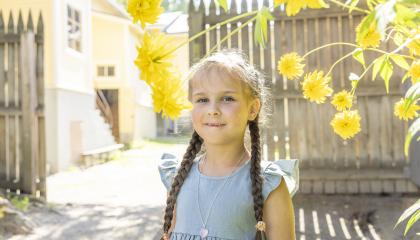 Pikkutyttö hymyilee kameralle, keltaisten kukkien joukosta. Taustalla museokorttelin taloja ja aitaa.