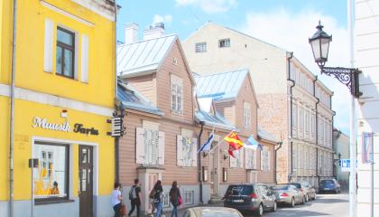 Vierekkäin kiinni toisissaan kaksi vanhaa rakennusta toinen keltainen kivitalo ja toinen vaalean ruskea vanha puutalo, joka on Tampere Maja nimeltään. Kadun varessa autoja ja ihmisiä kävelemässä kesäisessä säässä.