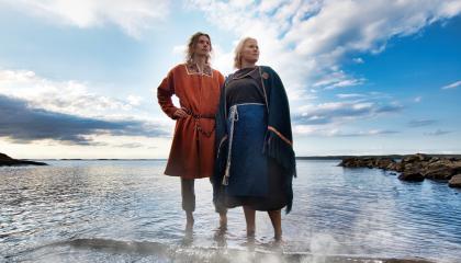 Mies ja nainen seisovat matalassa vedässä rannalla, taustalla sinistä taivasta ja pilviä. Vedästä nousee höyryä.