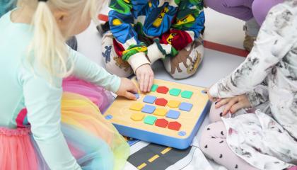 Kolme lasta istuu lattialla ja pelaa lautapeliä, jossa on värikkäitä nappuloita.