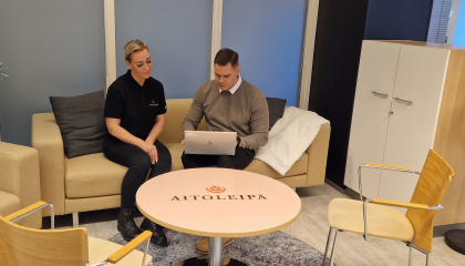 Kaksi henkilöä istuu sohvalla vieräkkäin. Oikeanpuoleiselle henkilöllä on kannettava tietokone sylissään ja hän esittelee vierustoverilleen näytöllä olevaa sisältöä. Huone on yrityksen taukotila, jossa sohva, kaappi ja nojatuoleja.