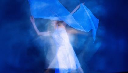 Valkoasuinen epätarkka hahmo tanssii vaalean kankaan kanssa sinistä taustaa vasten.