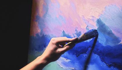 Käsi pitelee pensseliä, joka maalaa sinisiä ja vaaleanpunaisia sävyjä kankaalle.