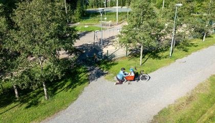 Vanhempi ja lapset leikkipuiston ulkopuolella pyörällä.