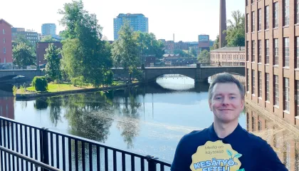 Tampereen kaupungin Kesätyösetelivastaava Petri Räsänen seisoo Vapriikinraitilla. Hän pitelee Kesätyösetelin logo ja seteleitä kädessään ja hymyilee. Taustalla näkyy Tampereen keskustaa.