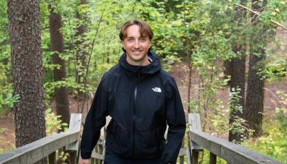 Henkilö seisoo hymyillen puuportaiden yläpäässä vehreän metsämaiseman keskellä.