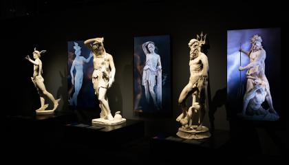 Kolme antiikin veistosta hämärässä näyttelytilassa valaistuna. Jokaisen taustalla videonäytöt.