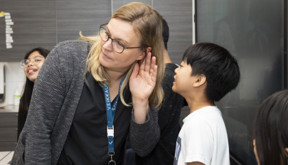 Lapsi kuiskaa opettajan korvaan. Taustalla toinen lapsi.