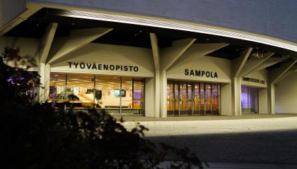 Sampola-rakennus pimeällä. Seinä on valaistu. Etualalla on pensas.