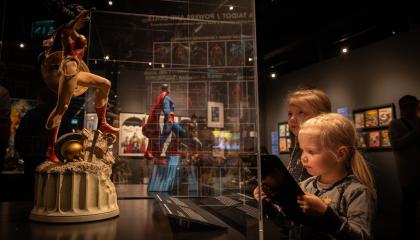 Hämärässä näyttelytilassa kaksi lasta katsoo vitriinissä olevaa Teräsmies ja Ihmenainen figuureja. 