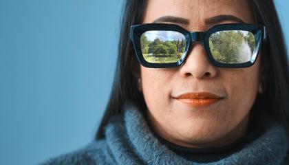 Tummahiuksinen henkilö katsoo suoraan kohti katsojaa. Hänellä on paksusankaiset aurinkolasit, joista heijastuu puiston kuva.