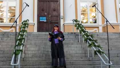 Nainen seisoo portailla ja lukee käsissään pitelemästään kansiosta runoa.