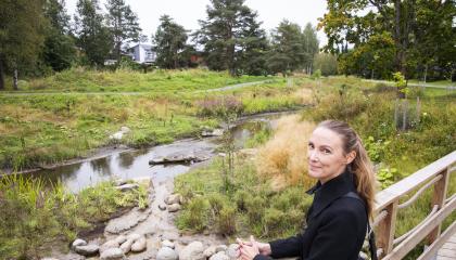 Tummatakkinen henkilö seisoo sillalla ihaillen ojaa, joka sijaitsee kauniissa ympäristössä.