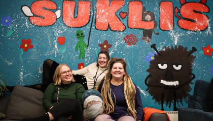 Kolme henkilöä istuu sohvalla ja hymyilee. Takana on seinä, jossa on Haisulin kuva ja jossa lukee Suklis.